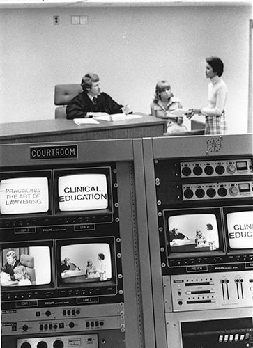A bank of monitors and other video equipment in the foreground and three individuals in the background talking with one another.